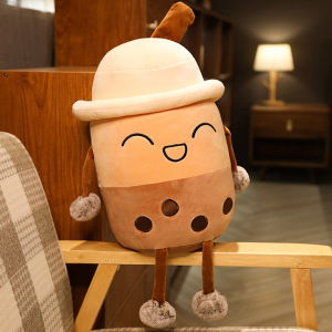 Pluszowa herbata bąbelkowa śmieje się w kapeluszu, jest beżowo-brązowa i siedzi na drewnianym podłokietniku fotela w beżowo-biało-brązową kratkę w mieszkaniu z zapaloną lampą w tle
