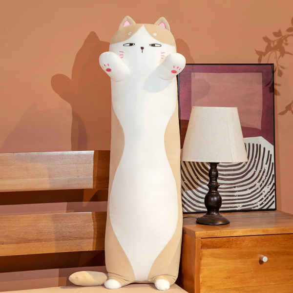 Na łóżku, obok którego stoi szafka nocna z białą lampką nocną i ramką za nią, znajduje się pluszowa biało-brązowa poduszka z kotem, stojącym na swoich malutkich tylnych łapkach
